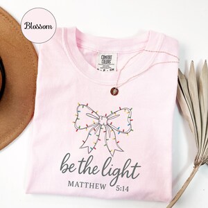 May include: A light pink t-shirt with a bow design made of Christmas lights. The text "be the light" and "Matthew 5:14" are printed below the bow. A rose gold necklace is draped over the shirt.