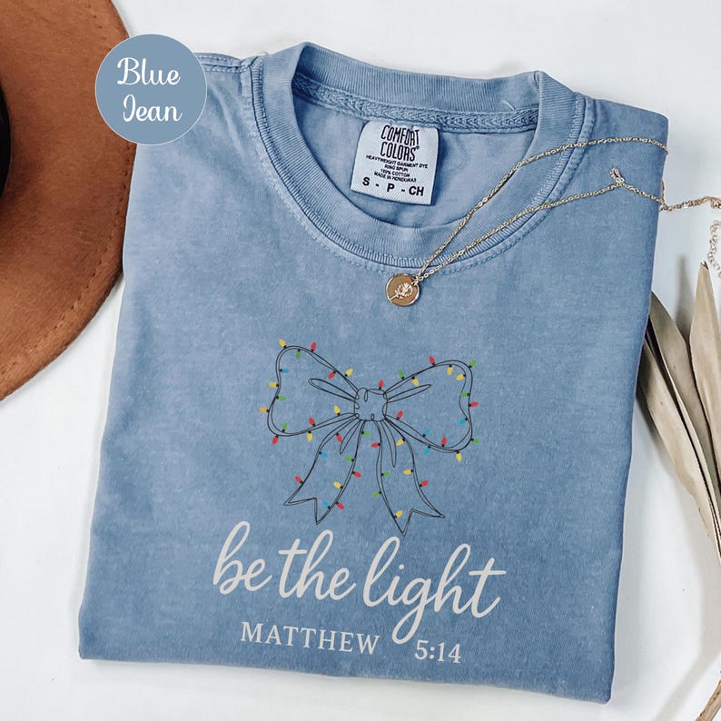 May include: Blue jean colored t-shirt with a bow design outlined in black and decorated with colorful lights. The words "be the light" and "Matthew 5:14" are printed in white. A gold necklace with a pendant is draped across the shirt.