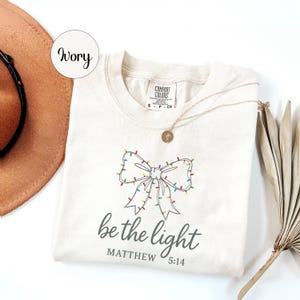 May include: Ivory-colored t-shirt with a bow design adorned with colorful Christmas lights. The text "be the light" and "Matthew 5:14" are printed below the bow. A gold necklace and a brown hat are also visible.