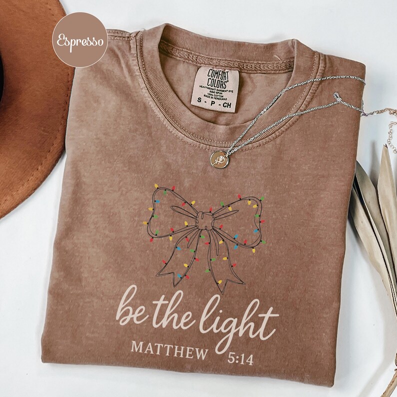 May include: Espresso-colored t-shirt with a bow design made of Christmas lights. The text "be the light" and "Matthew 5:14" are printed below the bow. A silver necklace with a pendant is draped across the shirt.