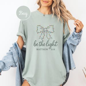 May include: A light green t-shirt with a bow design made of Christmas lights. The text "be the light" and "Matthew 5:14" are printed below the bow. The shirt is a relaxed fit.