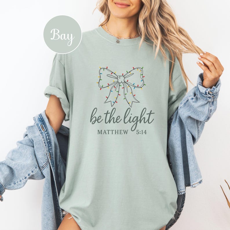 May include: A light green t-shirt with a bow design made of Christmas lights. The text "be the light" and "Matthew 5:14" are printed below the bow. The shirt is a relaxed fit.