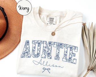 May include: An ivory t-shirt featuring the word AUNTIE in blue floral block letters, with the name Allison written in cursive below and a small bow icon. The shirt has a Comfort Colors tag at the neck and is displayed with a hat and dried leaves.