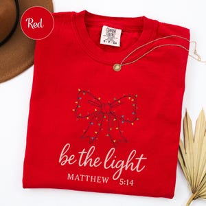 May include: A red crew neck t-shirt with a bow design made of Christmas lights. The text "be the light" and "Matthew 5:14" are printed in white. A gold necklace is draped across the shirt.