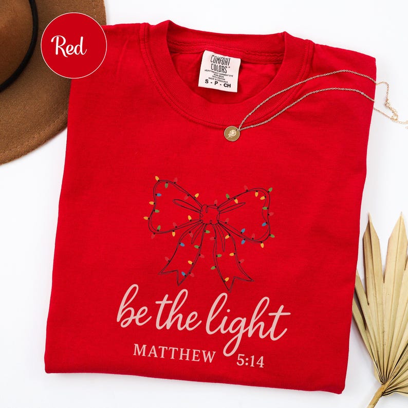 May include: A red crew neck t-shirt with a bow design made of Christmas lights. The text "be the light" and "Matthew 5:14" are printed in white. A gold necklace is draped across the shirt.