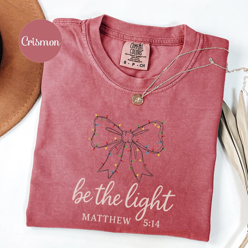May include: Crimson-colored t-shirt with a bow design outlined in black, adorned with colorful lights. The text "be the light" and "Matthew 5:14" are printed below the bow. A silver necklace with a pendant is draped on the shirt.