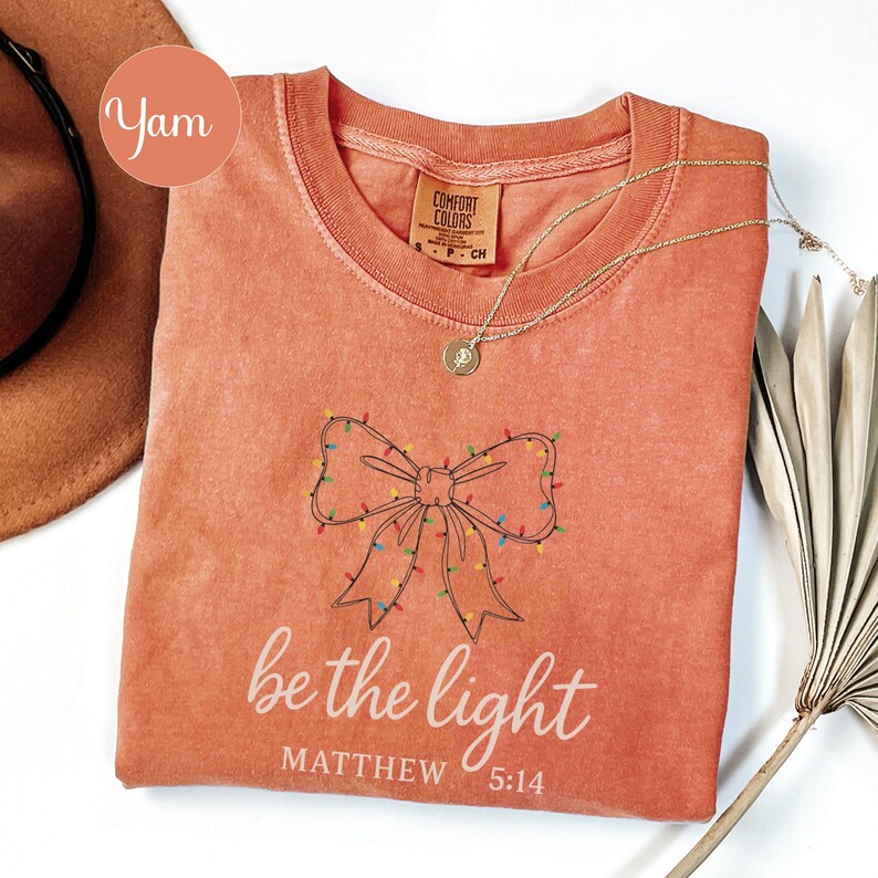 May include: Coral-colored t-shirt with a bow design adorned with colorful lights. The text "be the light" and "Matthew 5:14" are printed below the bow. A gold necklace with a pendant is draped on the shirt. The shirt is a Comfort Colors brand.