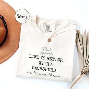 May include: Cream-coloured t-shirt with the text "LIFE IS BETTER WITH A DACHSHUND or two or three" in black. A small dachshund graphic is above the text. A gold necklace and a brown hat are also visible.