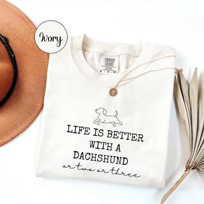 May include: Cream-coloured t-shirt with the text "LIFE IS BETTER WITH A DACHSHUND or two or three" in black. A small dachshund graphic is above the text. A gold necklace and a brown hat are also visible.