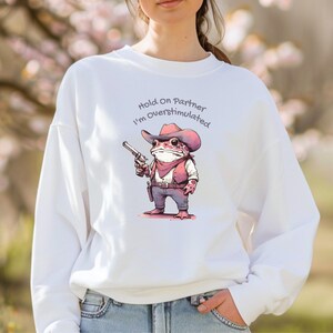 May include: White sweatshirt featuring a cartoon frog dressed as a cowboy, holding a gun. The frog wears a hat, vest, and bandana. Text above the frog reads "Hold On Partner I'm Overstimulated."
