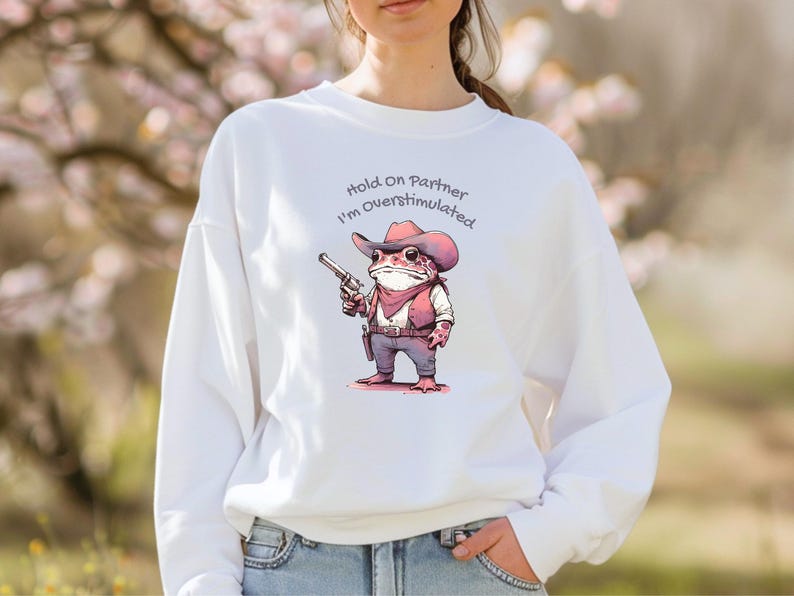 May include: White sweatshirt featuring a cartoon frog dressed as a cowboy, holding a gun. The frog wears a hat, vest, and bandana. Text above the frog reads "Hold On Partner I'm Overstimulated."