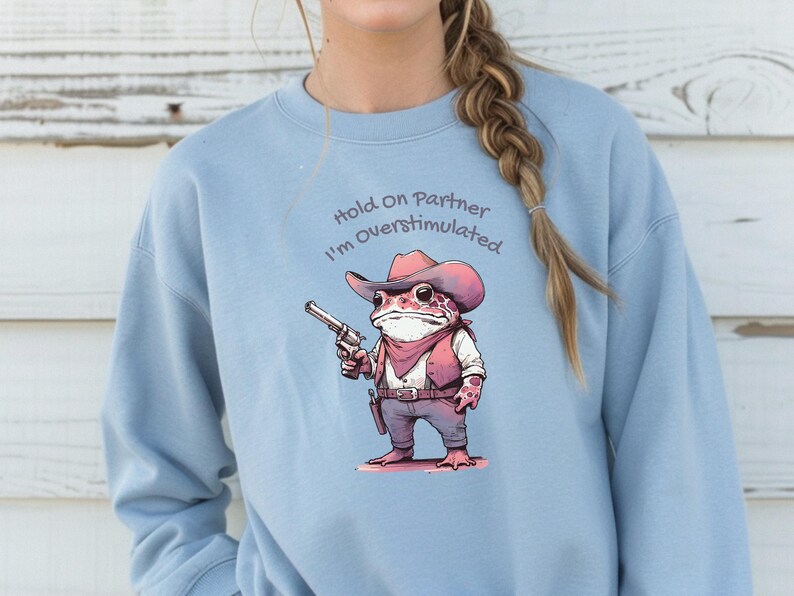 May include: Light blue sweatshirt featuring a cartoon frog dressed as a cowboy, holding a gun. The frog wears a pink cowboy hat, vest, and bandana. Text above the frog reads "Hold On Partner I'm Overstimulated."