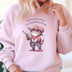 May include: Light pink sweatshirt featuring a cartoon frog dressed as a cowboy, holding a gun. The frog wears a cowboy hat, vest, and bandana. Text above the frog reads "Hold On Partner I'm Overstimulated."