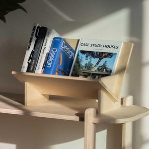 May include: A light-colored wooden bookshelf with several books. The books include titles like "Design" and "Case Study Houses." A clear glass vase with a large green leaf is in the background. The bookshelf is on a light-colored shelf.