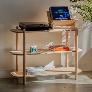 May include: A light-colored wooden record player stand with three shelves. The top shelf holds a black record player and a record holder with a record that says "April Orchestra FR2". The middle shelf has books and a small camera. The bottom shelf has books.