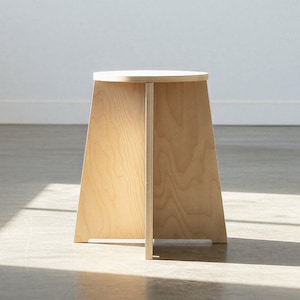 Op de afbeelding: Een minimalistische houten kruk met een ronde bovenkant en een trapeziumvormige basis. De kruk is gemaakt van lichtgekleurd hout en heeft een eenvoudig, modern ontwerp. De kruk is ongeveer 46 cm hoog.