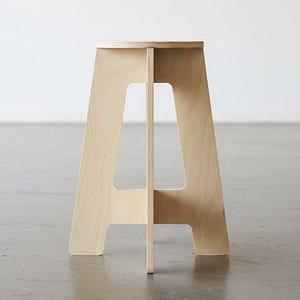 May include: A light-colored wooden stool with an A-frame design. The stool has a flat, round seat and a sturdy base. The wood grain is visible, and the stool is set against a white wall and a gray floor.