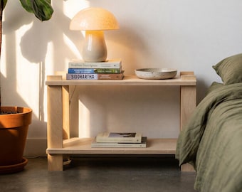 May include: A light wood bedside table with a mushroom-shaped lamp, books, and a speckled bowl. A potted plant is to the left, and a bed with olive green bedding is on the right. The lamp emits a warm glow.