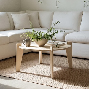 May include: A light-colored, sectional sofa with white cushions and pillows. A round, light wood coffee table holds a bowl of greenery, a mug, and a book. A woven rug and framed botanical prints complete the neutral-toned living space.