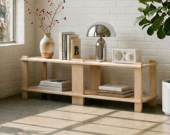 May include: A light-colored wooden shelf unit with two levels, holding books, a chrome lamp, and a small white speaker. A vase with red stems and a potted plant are also present. The unit is against a white brick wall.