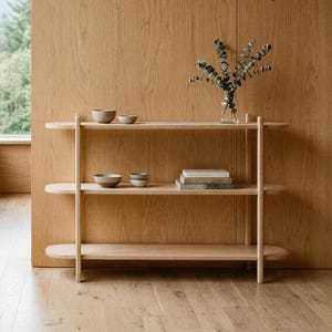 May include: A light-colored wooden shelving unit with three rounded shelves supported by two vertical poles. The top shelf holds two small bowls, the middle shelf holds two small bowls and a stack of books, and the top shelf holds a glass vase with greenery.