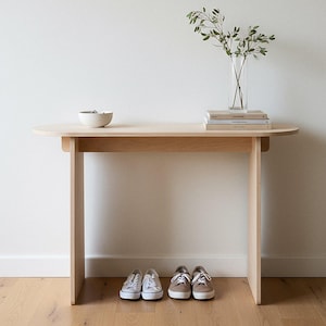 May include: A light-colored wooden console table with a rounded top. A small white bowl and a clear vase with greenery sit on the table. Two pairs of sneakers are placed beneath the table.
