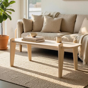 May include: A light wood coffee table with rounded edges and angled legs sits on a textured beige rug. A beige sofa with matching pillows and a blanket is in the background. A potted plant and a mug are also visible.