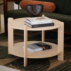 Peut inclure: Une table basse ronde &agrave; deux niveaux en bois clair, avec une finition naturelle. L'&eacute;tag&egrave;re sup&eacute;rieure contient deux livres et un bol en m&eacute;tal fonc&eacute;. L'&eacute;tag&egrave;re inf&eacute;rieure a un seul livre. La table est devant un canap&eacute; vert.