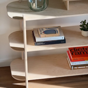 May include: A light wood bookshelf with three shelves. The shelves are rounded at the front. The top shelf has a clear glass vase with a green plant. The middle shelf has a stack of books, including "The Bugatti Story" and "Kinfolk". The bottom shelf has a stack of books with the title "Dieter Rams".
