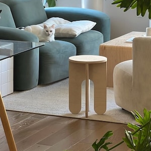 May include: Two white cats relaxing on a teal blue sofa with a white throw pillow. A wooden stool with rounded legs sits in front of the sofa. A large potted plant with green leaves is in the background.