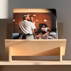 May include: A wooden shelf unit with a framed photograph and a stack of books. The photograph depicts a family scene. The books include "The Kinfolk Entrepreneur" and "Axel Vervoordt Portraits of Interiors". The shelf is made of light-colored wood.