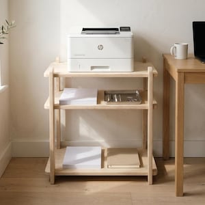May include: A light-colored wooden shelving unit with three shelves. A white printer sits on the top shelf. The middle shelf holds a metal tray and stacks of white paper. A laptop and mug are on a wooden desk.