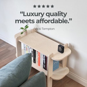 May include: A light-colored wooden bookshelf with rounded edges and multiple shelves. The top shelf holds a small plant and a black clock. The bookshelf is filled with books of various colors. Text on the image reads: "Luxury quality meets affordable."