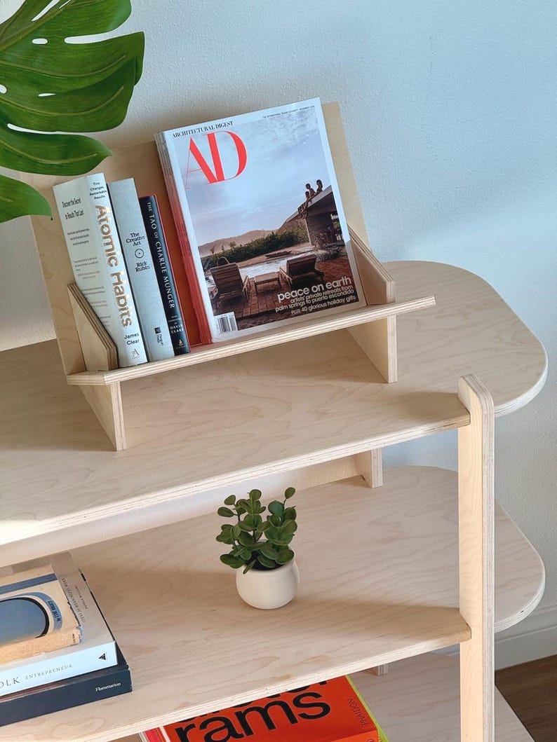 May include: A light-colored wooden bookshelf with multiple shelves. The top shelf holds a magazine with a red "AD" logo and several books, including "Atomic Habits." A small potted plant sits on a lower shelf.