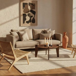 May include: A living room scene featuring a beige sofa with several throw pillows, a round wooden coffee table, and a woven chair. A framed abstract art piece hangs on the wall. Candles and a large terracotta vase add decorative elements.