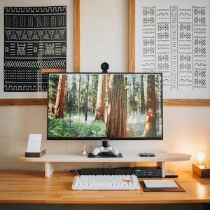 May include: A white keyboard with a red key on a wooden desk with a monitor displaying a forest scene. The monitor is on a wooden stand with a black video game controller on top. There are two framed black and white patterned prints on the wall behind the desk.