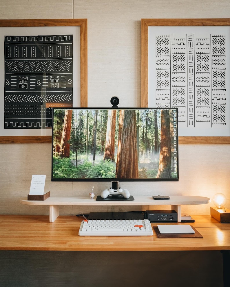 May include: A white keyboard with a red key on a wooden desk with a monitor displaying a forest scene. The monitor is on a wooden stand with a black video game controller on top. There are two framed black and white patterned prints on the wall behind the desk.