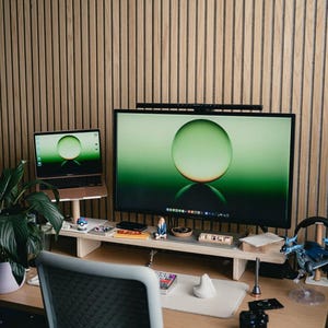 May include: A home office workspace with a wooden desk, a laptop, a large monitor, a plant, and a chair. The monitor is displaying a green sphere on a green background. The desk is organized with various items, including a small figurine, a keyboard, and a mouse.