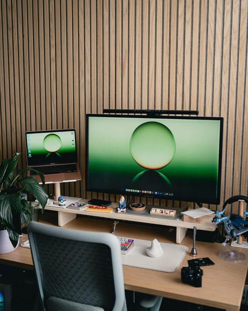 May include: A home office workspace with a wooden desk, a laptop, a large monitor, a plant, and a chair. The monitor is displaying a green sphere on a green background. The desk is organized with various items, including a small figurine, a keyboard, and a mouse.