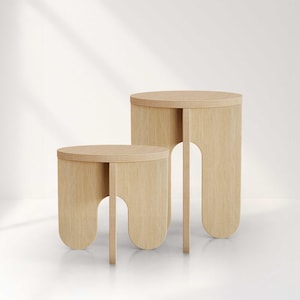 May include: Two light-colored wooden side tables. The tables have a round top and a unique leg design with a curved arch. The smaller table is in front of the larger one. The wood grain is visible, giving a natural look.