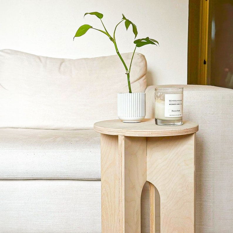 Op de afbeelding: Een lichte houten bijzettafel met een rond blad en drie poten. Een witte keramische plantenpot met een groene plant en een glazen kaarsglas staan op de tafel.