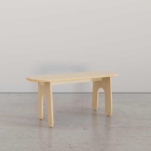 Puede incluir: Un banco de madera clara con la parte superior redondeada y patas curvas &uacute;nicas. El banco est&aacute; sobre un suelo de hormig&oacute;n.