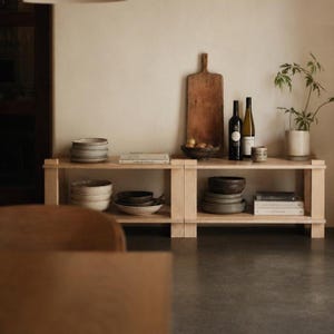 May include: A light wood, low-profile shelving unit displays various kitchen and dining items. The shelves hold stacks of plates, bowls, books, wine bottles, a cutting board, and a small potted plant. The items are in neutral tones.