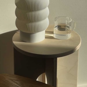Op de afbeelding: Een witte keramische lamp met een golvend ontwerp staat op een licht houten bijzettafel met een glas water. De tafel heeft een afgerond blad en een gebogen, houten onderstel.