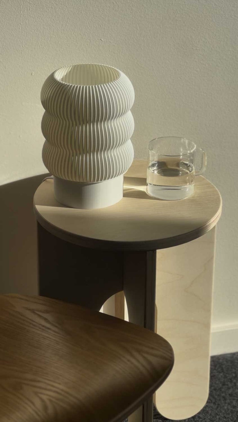 Op de afbeelding: Een witte keramische lamp met een golvend ontwerp staat op een licht houten bijzettafel met een glas water. De tafel heeft een afgerond blad en een gebogen, houten onderstel.