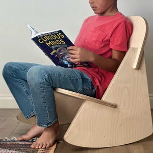 May include: A child sits in a light wood rocking chair reading a book titled "Interesting Facts for Curious Minds". The child is wearing blue jeans and a red t-shirt.