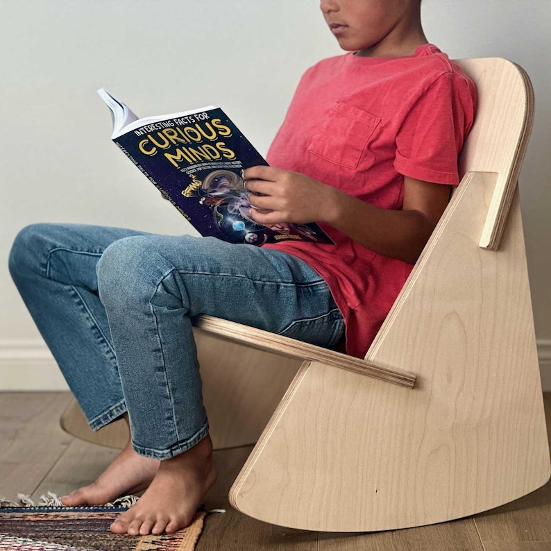 May include: A child sits in a light wood rocking chair reading a book titled "Interesting Facts for Curious Minds". The child is wearing blue jeans and a red t-shirt.
