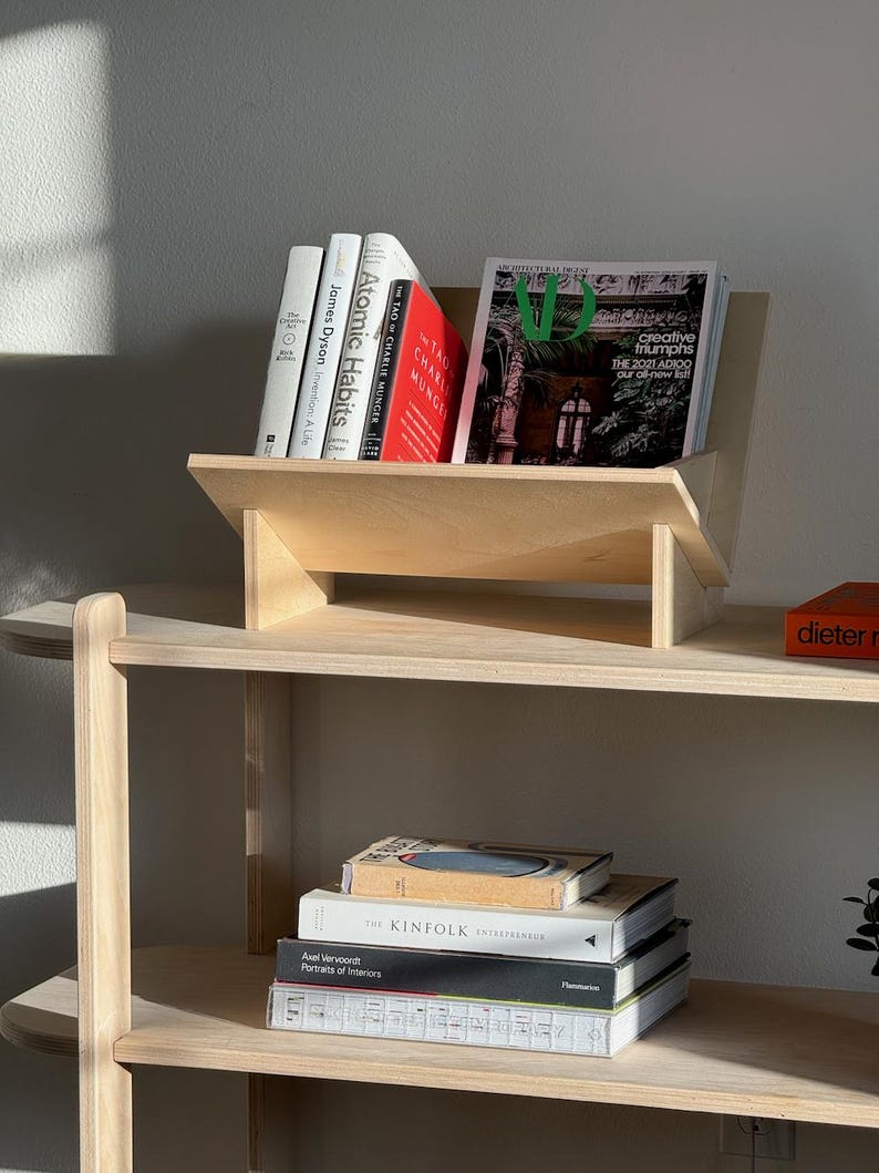 May include: A wooden bookshelf with a built-in angled book display. Several books are visible, including "Atomic Habits" and "The Tao of Charlie Munger." A magazine with the text "creative triumphs" is also displayed. Additional books are stacked on a lower shelf.