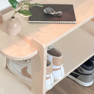 May include: A light-coloured wooden shoe rack with three shelves. The top shelf holds a small vase with a plant, a watch, and a notebook. The lower shelves display pairs of trainers in white, black, and grey.