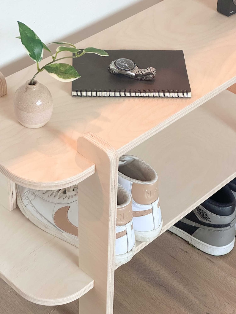 May include: A light-coloured wooden shoe rack with three shelves. The top shelf holds a small vase with a plant, a watch, and a notebook. The lower shelves display pairs of trainers in white, black, and grey.